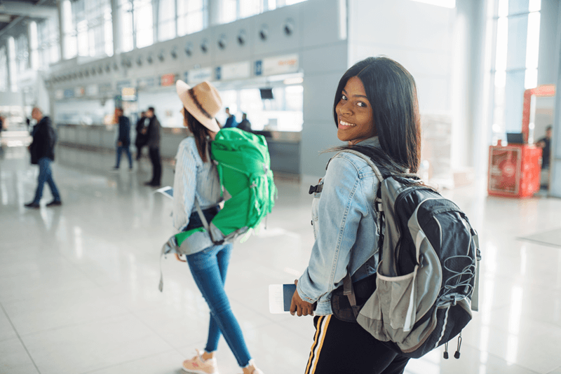 Jovens viajantes com mochilas caminhando em aeroporto, representando intercâmbio internacional e experiência de estudo no exterior.