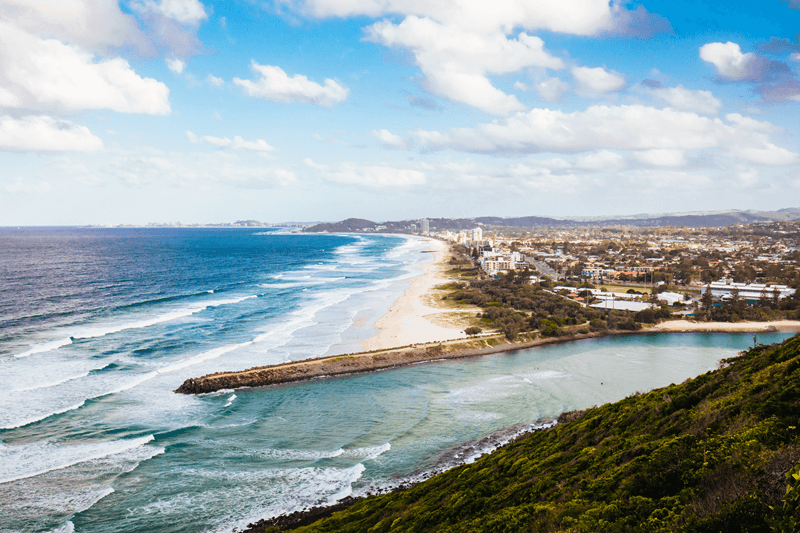 Vista aérea de praia na Austrália com mar azul e cidade ao fundo, destacando o estilo de vida de quem estuda e trabalha no país.