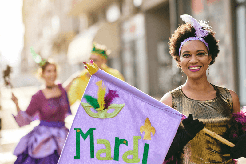 Participante do Mardi Gras vestindo fantasia roxa e dourada segurando bandeira decorada durante celebração de Carnaval em Nova Orleans.